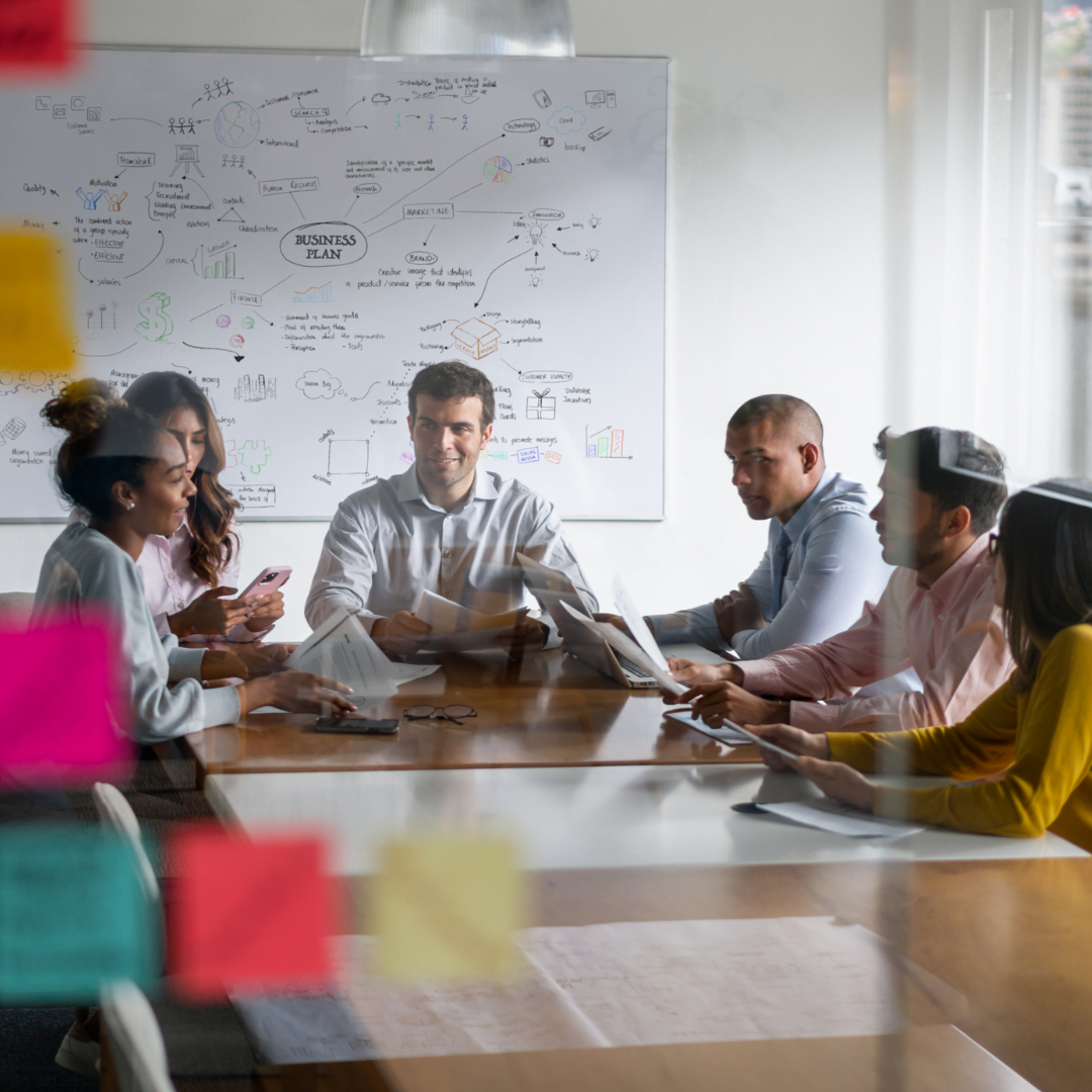 People-around-a-board-room-table-discussing-a-plan-brainstormed-on-board-behind-them